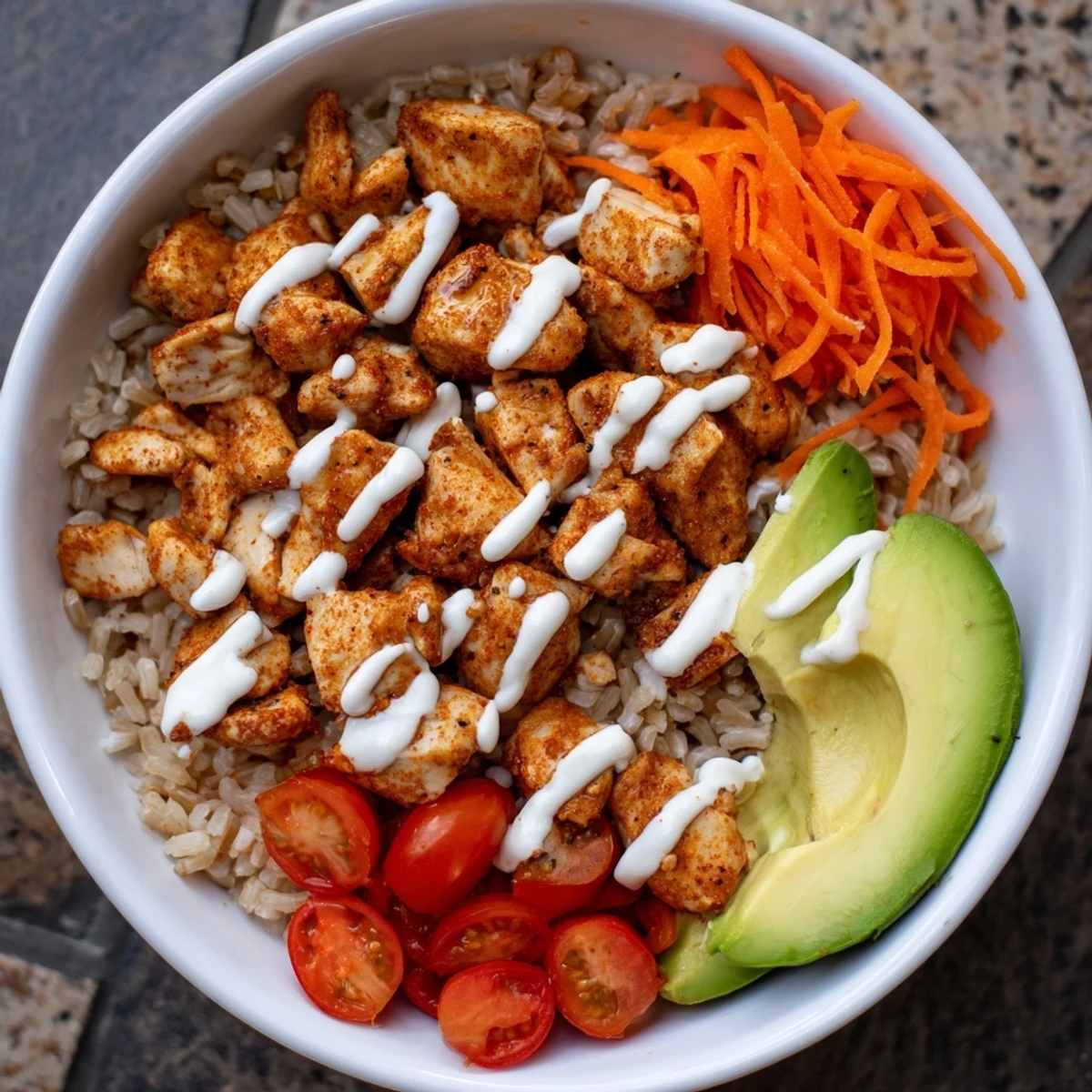 A close-up of a Spicy Lunch Bowl with tender chicken, brown rice, avocado, and vibrant fresh vegetables, drizzled with creamy red sauce.