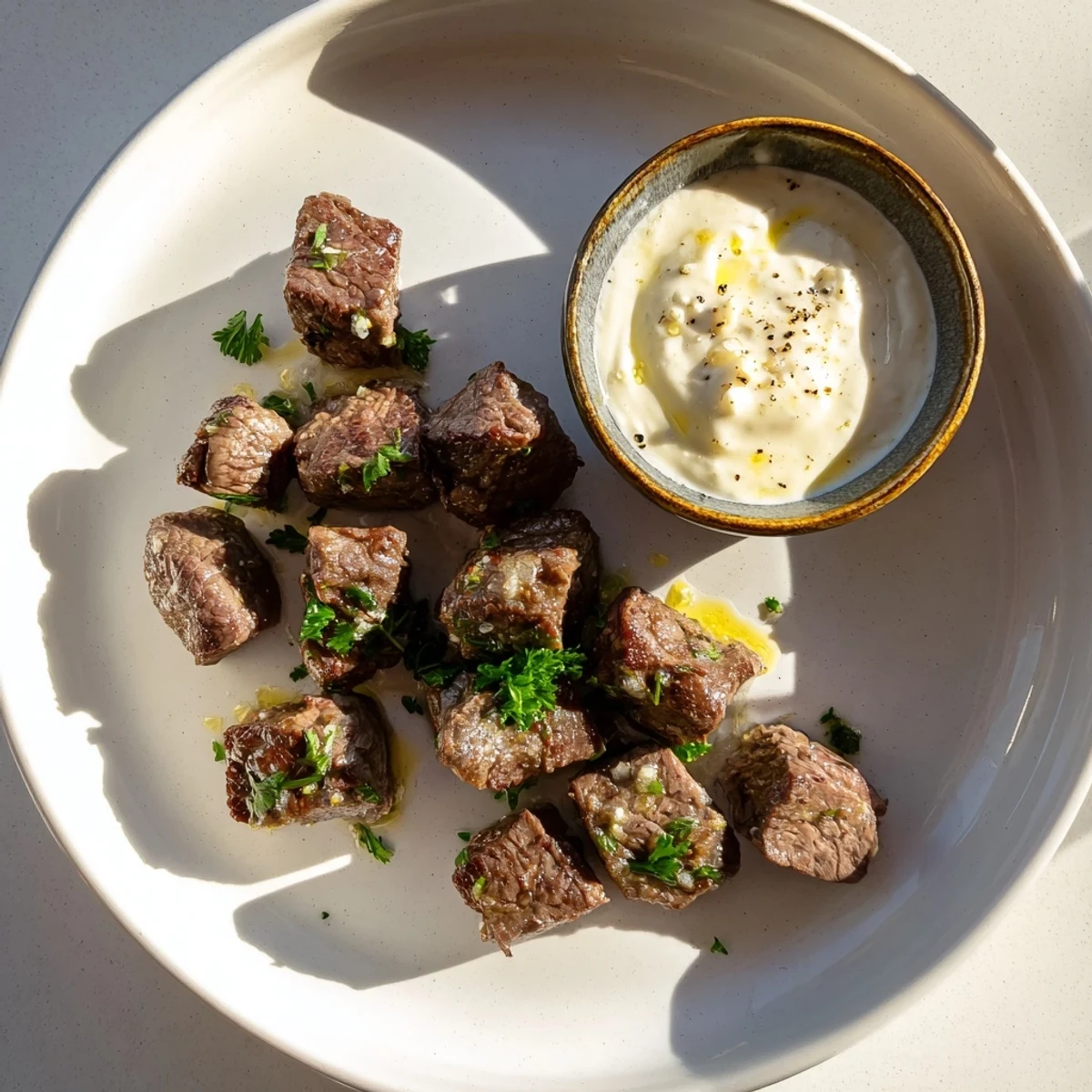 Golden-brown garlic butter steak bites glistening on a platter with a creamy white horseradish dip, garnished with fresh parsley.