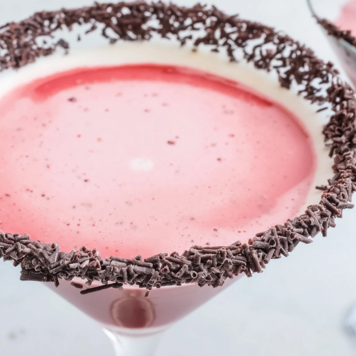 A close-up of a Decadent Red Velvet Martini in a chilled glass, rimmed with cream cheese frosting and chocolate shavings, garnished with crumbs.